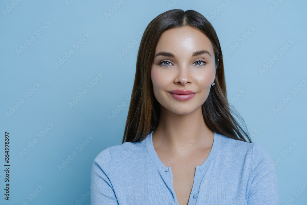 Beautiful brunette young woman has long dark hair, poses indoor against blue studio background, being successful businesswoman, has natural look, enjoys leisure or day off, stands delighted. Beautiful brunette young woman has long dark hair, poses indoor against blue studio background, being successful businesswoman, has natural look, enjoys leisure or day off, stands delighted.