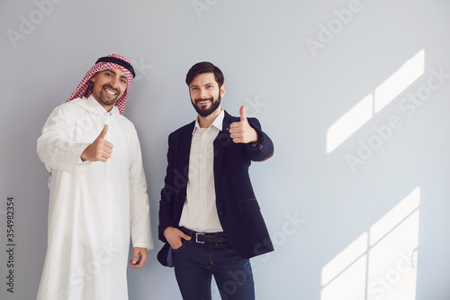 Arab and european businessmen show thumb up stand smiling at the gray background.