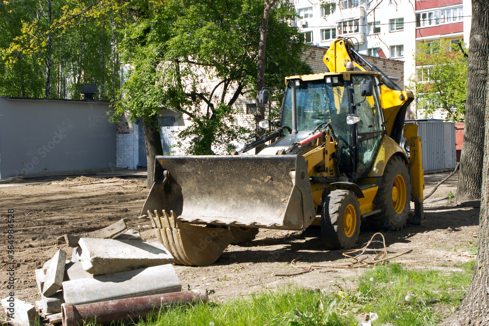 The yellow bulldozer excavator is located in the courtyard of a multi-storey multi-apartment building. Replacement of heating networks in the doorway of the house, overhaul. The yellow bulldozer excavator is located in the courtyard of a multi-storey multi-apartment building. Replacement of heating networks in the doorway of the house, overhaul.