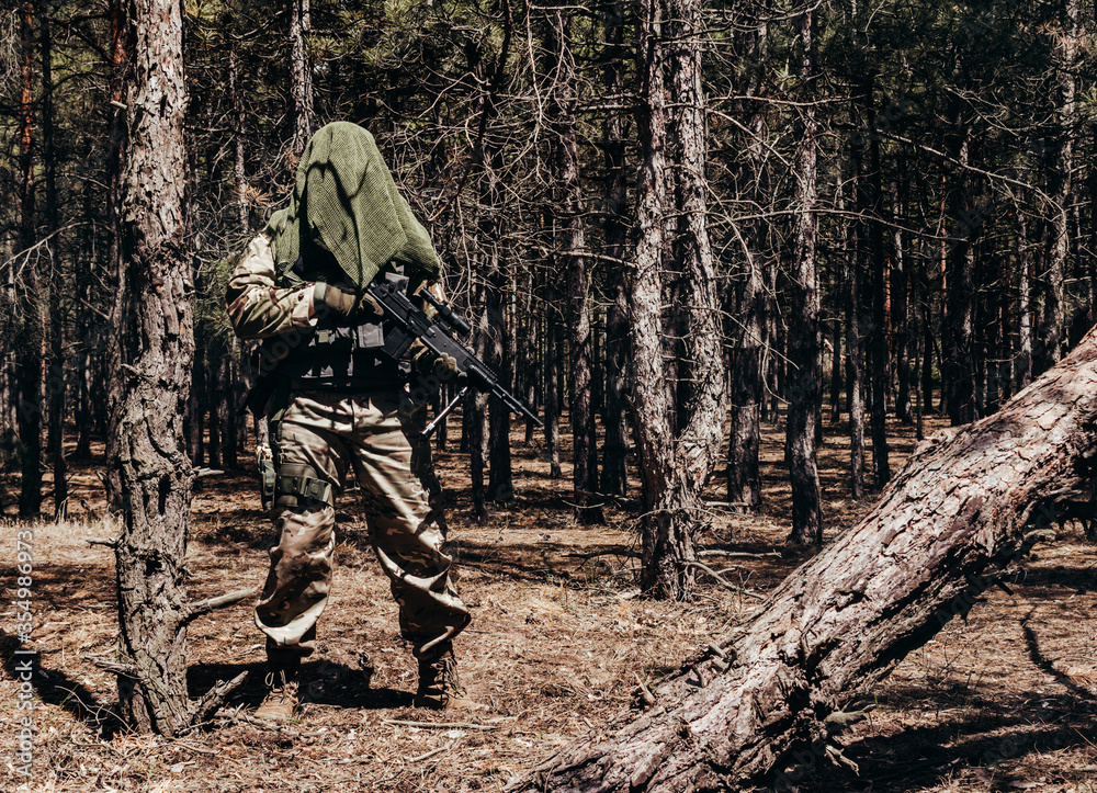 Forest sniper in camouflage, net scarf and armor vest standing with rifle.