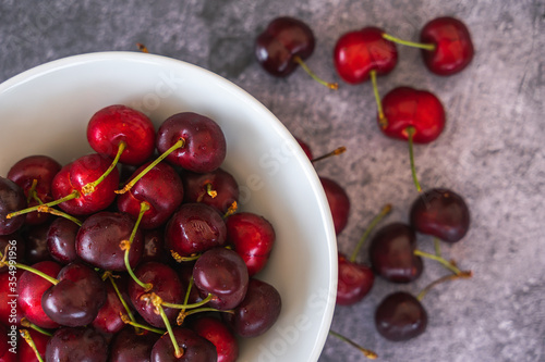 Top view of fresh ripe cherries in a white bowl on a slate surface with a few more cherries scattered on it.