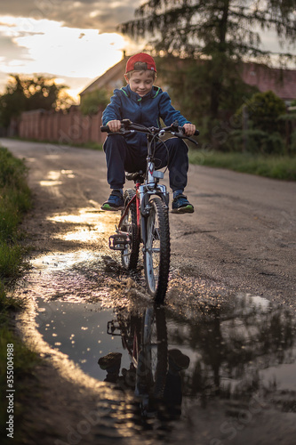 Young boy rides a bicycle through a puddle on a dirt road.