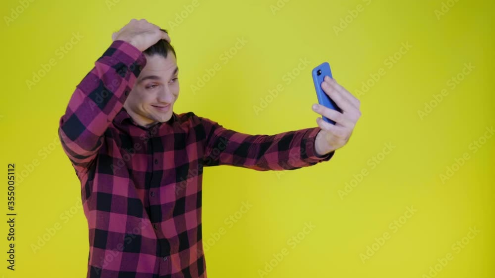 Hello or bye. Cheerful male blogger in a pink plaid shirt is recording video on the smartphones selfie camera for subscribers on social networks on a yellow background with copy space Hello or bye. Cheerful male blogger in a pink plaid shirt is recording video on the smartphones selfie camera for subscribers on social networks on a yellow background with copy space