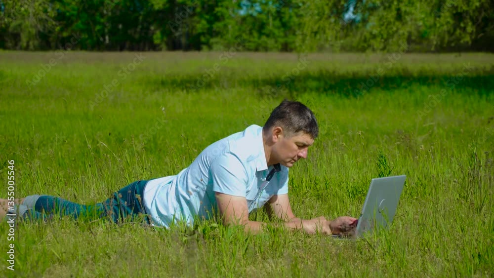 Man Works behind a Laptop lying on the Grass At The Sunny Day. Freelancer Remote Work Concept. 4K Man Works behind a Laptop lying on the Grass At The Sunny Day. Freelancer Remote Work Concept. 4K