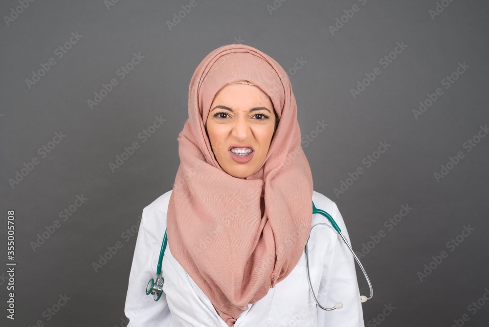 Mad crazy doctor woman clenches teeth angrily, being annoyed with coming noise, dressed in medical uniform, isolated over gray background. Negative feeling concept. Mad crazy doctor woman clenches teeth angrily, being annoyed with coming noise, dressed in medical uniform, isolated over gray background. Negative feeling concept.