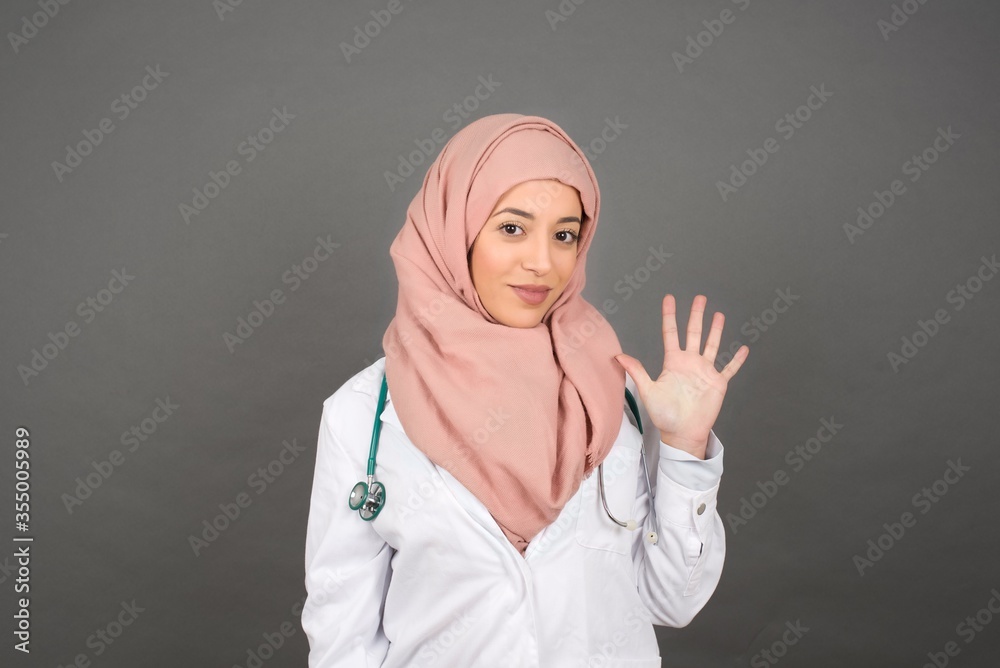 Young muslim doctor woman standing against gray wall showing and pointing up with fingers number five while smiling confident and happy. Young muslim doctor woman standing against gray wall showing and pointing up with fingers number five while smiling confident and happy.