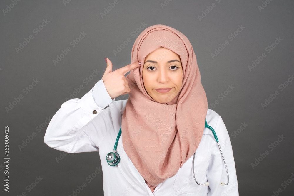 Unhappy muslim doctor woman curves lips and makes suicide gesture, tired of everything, shoots in temple with hand, isolated against studio background. Unhappy muslim doctor woman curves lips and makes suicide gesture, tired of everything, shoots in temple with hand, isolated against studio background.