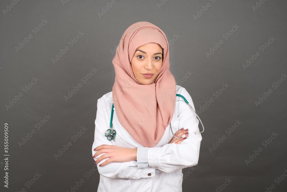 Waist up portrait of beautiful young doctor muslim female with positive expression, has crossed arms, feels happy and confident, wears casual clothes, isolated over gray background Waist up portrait of beautiful young doctor muslim female with positive expression, has crossed arms, feels happy and confident, wears casual clothes, isolated over gray background