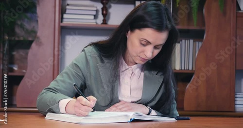 Brunette attractive woman writing in a white large notebook or journal and looks to phone. Education, business, working from home concept. Medium shot. photo