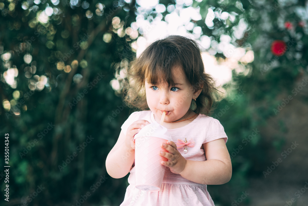 A beautiful little girl in a pink dress against a background of plants drinks a drink from a cocktail glass A beautiful little girl in a pink dress against a background of plants drinks a drink from a cocktail glass
