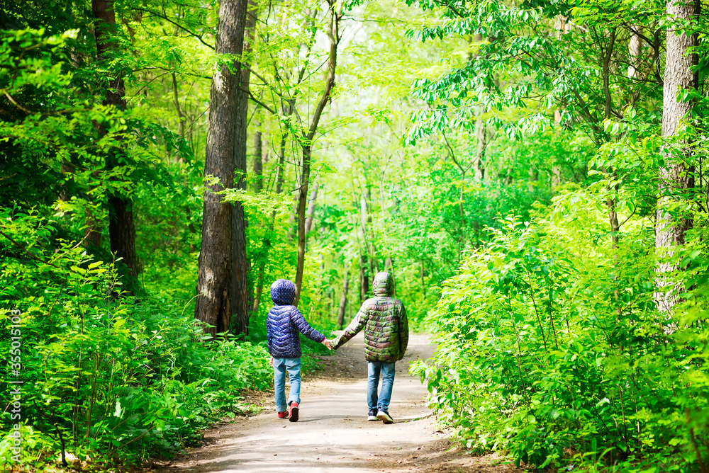 Two children in jackets are walking around city park or forest on a safe distance in a sunny day holding hands Two children in jackets are walking around city park or forest on a safe distance in a sunny day holding hands