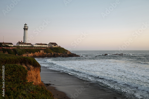 One old white ship lighthouse with buildings on the steep rocky coast of the raging ocean in the evening at sunset with beautiful sky and green grass waiting for ships