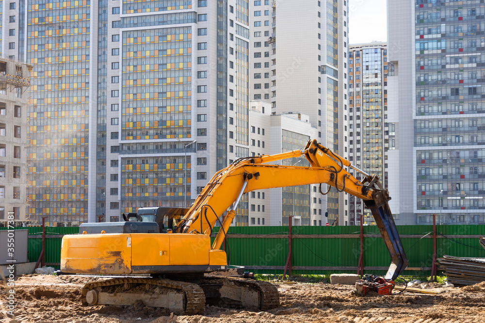 Obraz premium Heavy excavator in a working high frequency hydraulic vibratory mounted pile driver on road against the background of residential multi-storey buildings.