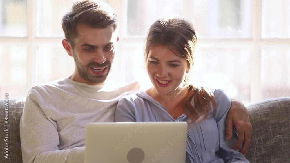Couple sitting on couch browse internet offers read media news, husband embraces wife resting at home spend time use laptop feels surprised happy about cheap flight tickets travel opportunity concept