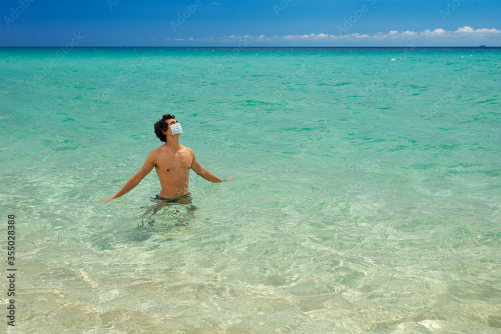Young handsome man bathing in paradise sea with surgery protective mask on a bright sunny day during 2020 coronavirus summer holiday Young handsome man bathing in paradise sea with surgery protective mask on a bright sunny day during 2020 coronavirus summer holiday