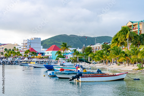 Boats docked on tropical Caribbean island coast with white sand and coconut palm trees. Tourists and locals shop at stores and restaurants on shore. Mountain landscape in background. photo