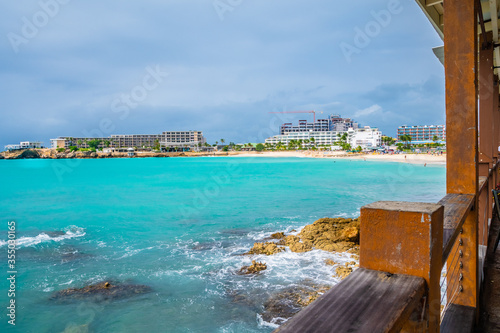 Scenic ocean view from traditional Caribbean seaside restaurant. Hotels and resorts on coastline of tropical island coast. People/ tourists on white sand beach shore.