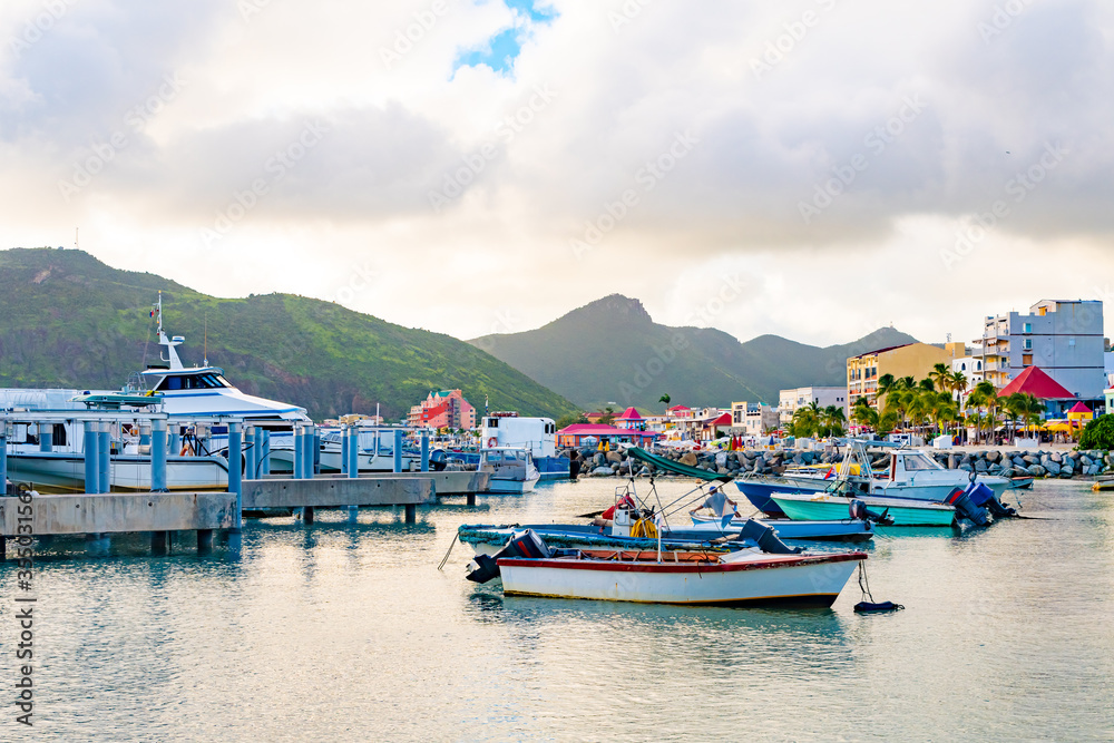 Motorboats, speedboats and yachts docked in boatyard on tropical Caribbean island coast. Coconut palm trees, colorful buildings with shops and restaurants in background. Mountain landscape in distance