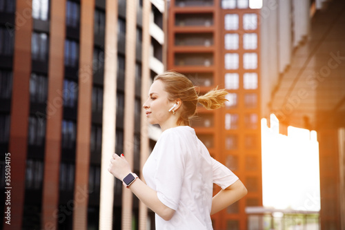 Fotografie Woman in white t shirt running on street of city near building and skyscrapers in wireless headphones with smart watch