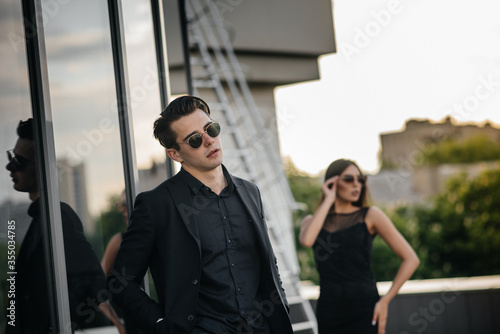 A beautiful, stylish pair of young people in black clothes and glasses stand against the background of an office building in the sunset. Fashion and style