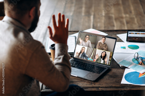 Video communication. Back view of a young man who communicates by video communication with his employees, he greets them waving his right hand. Distant communication. Work from home photo