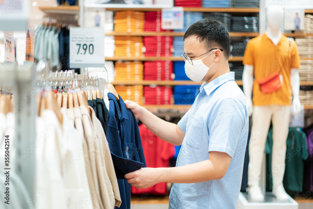 Asian man wearing mask over her face while choosing shirt at shopping mall with shopping bag for healthcare and prevention from coronavirus, Covid19 influenza in crowded place.. Asian man wearing mask over her face while choosing shirt at shopping mall with shopping bag for healthcare and prevention from coronavirus, Covid19 influenza in crowded place..