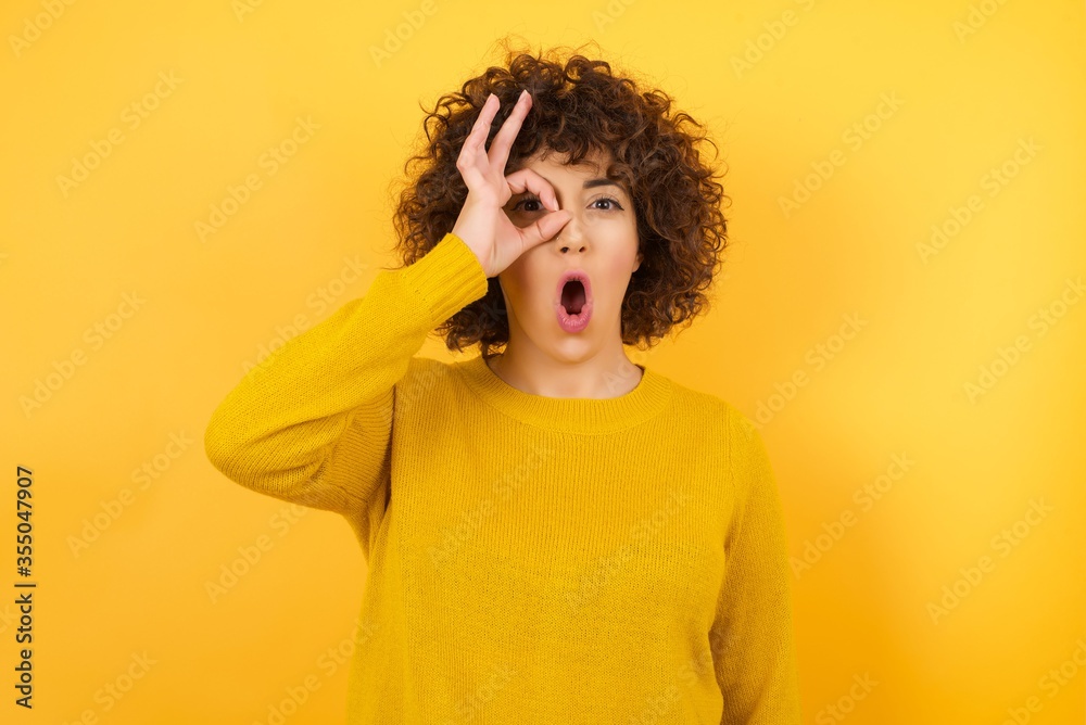 Young african american woman wearing yellow sweater standing over isolated yellow background doing ok gesture shocked with surprised face, eye looking through fingers. Unbelieving expression. Young african american woman wearing yellow sweater standing over isolated yellow background doing ok gesture shocked with surprised face, eye looking through fingers. Unbelieving expression.