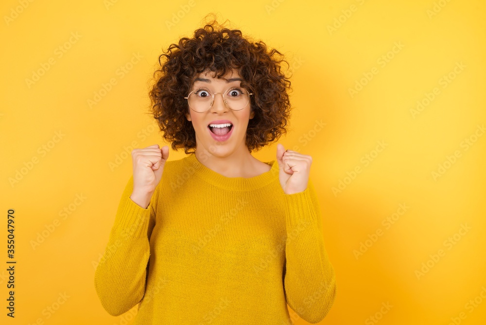 Young beautiful Arab businesswoman with curly hair wearing yellow sweater celebrating surprised and amazed for success with arms raised and open eyes. Winner concept. Young beautiful Arab businesswoman with curly hair wearing yellow sweater celebrating surprised and amazed for success with arms raised and open eyes. Winner concept.