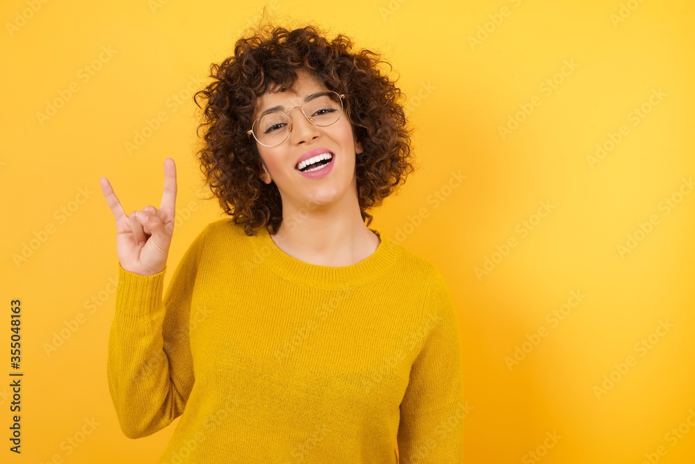 Young arab girl with blue eyes doing a rock gesture and smiling to the camera. Wearing yellow sweater standing against yellow wall. Ready to go to her favorite band concert. Young arab girl with blue eyes doing a rock gesture and smiling to the camera. Wearing yellow sweater standing against yellow wall. Ready to go to her favorite band concert.