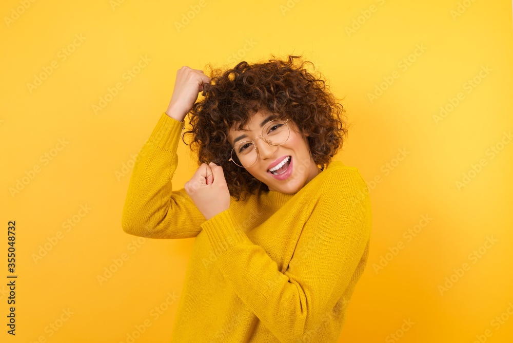 Profile photo of excited beautiful Arab businesswoman wearing yellow sweater and glasses raising fists celebrating black friday shopping wears yellow sweater isolated over yellow background Profile photo of excited beautiful Arab businesswoman wearing yellow sweater and glasses raising fists celebrating black friday shopping wears yellow sweater isolated over yellow background