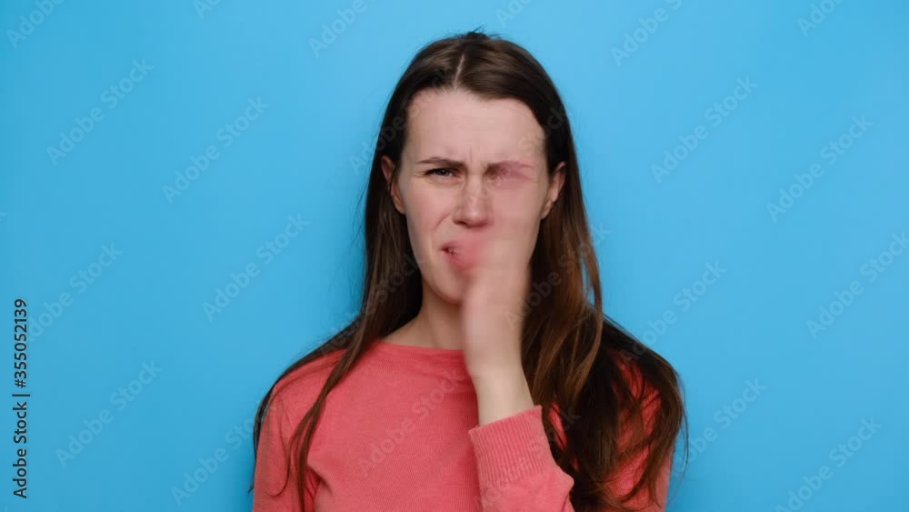 Close up portrait of annoyed young brunette woman checking her breath, smells something awful, pinches nose, dressed in sweater, isolated over blue studio background. Bad odour concept. Close up portrait of annoyed young brunette woman checking her breath, smells something awful, pinches nose, dressed in sweater, isolated over blue studio background. Bad odour concept.