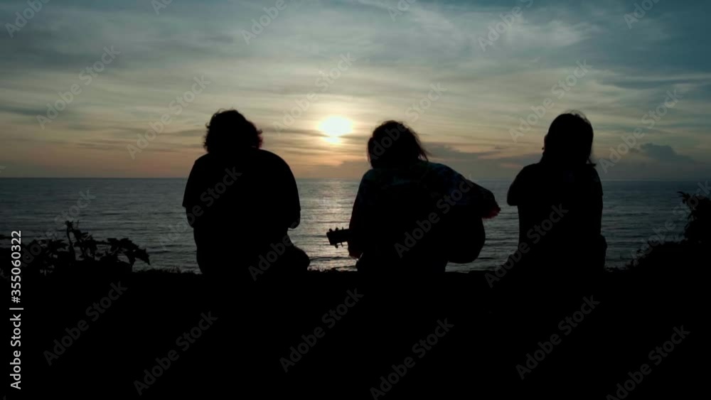 Silhouettes of three friends sitting on a mountain at sunset in front of the sea Silhouettes of three friends sitting on a mountain at sunset in front of the sea