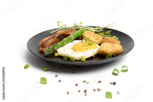 a fried egg sunny side up on a black plate with potatoes bacon asparagus and bread isolated on white
