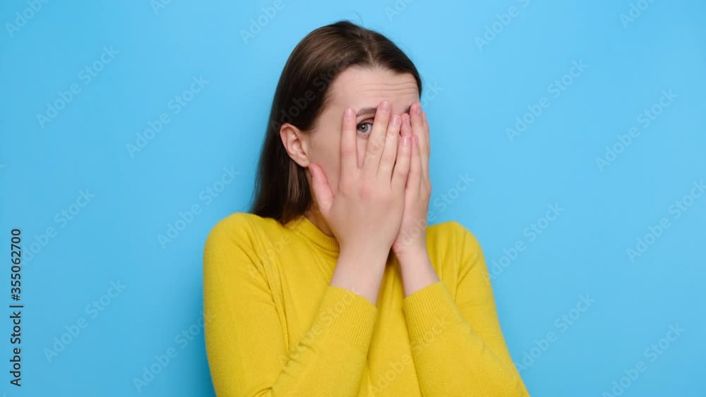 Portrait of scared young woman peep through fingers feel frightened terrified, stares with widely opened eyes, wears yellow sweater, hides face from someone, isolated on blue studio background Portrait of scared young woman peep through fingers feel frightened terrified, stares with widely opened eyes, wears yellow sweater, hides face from someone, isolated on blue studio background