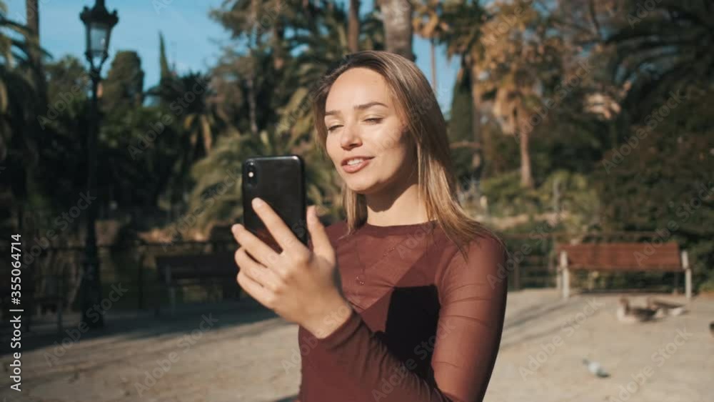 Tracking shot of pretty girl walking through city park and happily discussing news with family on video call on smartphone Tracking shot of pretty girl walking through city park and happily discussing news with family on video call on smartphone