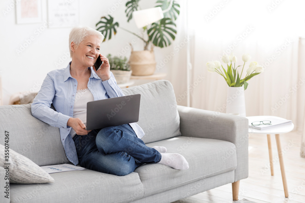 Happy senior lady sitting on couch, holding laptop and talking on cellphone Happy senior lady sitting on couch, holding laptop and talking on cellphone