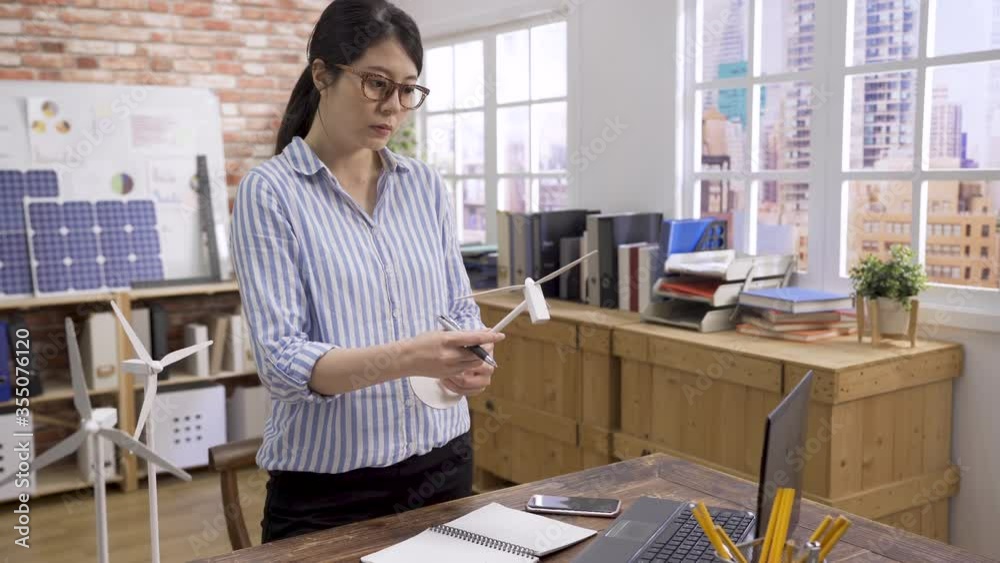 beautiful wind turbine engineer woman working in modern office. green marketing concept. young asian korean female architect examining 3D windmill model and writing standing at table in company. beautiful wind turbine engineer woman working in modern office. green marketing concept. young asian korean female architect examining 3D windmill model and writing standing at table in company.