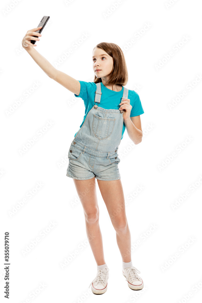 Teen girl in denim overalls and a blue T-shirt makes selfie on a smartphone. Blogging and online communication. Isolated on a white background. Full height. Vertical. Teen girl in denim overalls and a blue T-shirt makes selfie on a smartphone. Blogging and online communication. Isolated on a white background. Full height. Vertical.