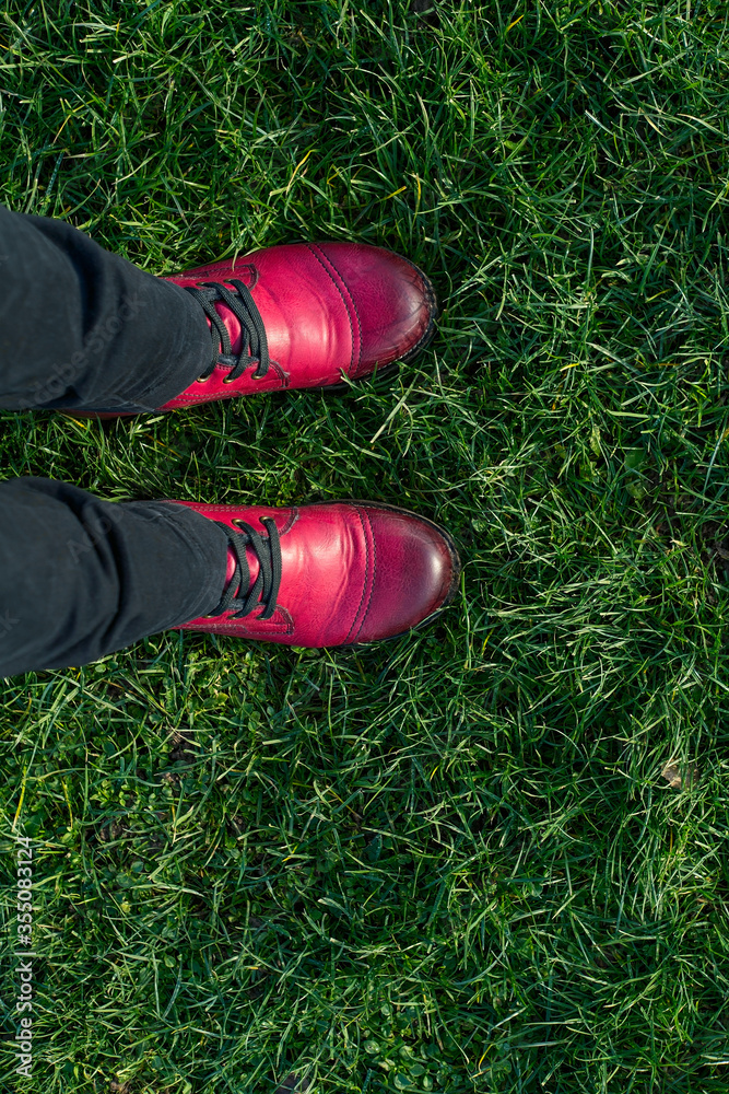 Fototapeta premium Feet of a woman in red leather shoes and trousers standing on the green grass, top view