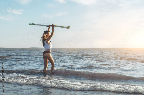 Young caucasian woman surfer in white shirt going to the sea with her surfboard above her head.