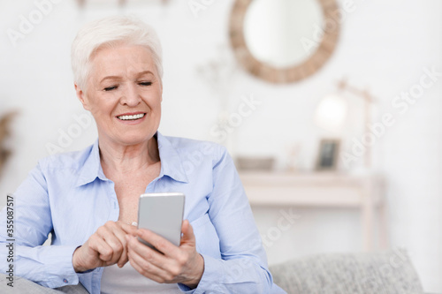 Smiling elderly woman holding cellphone, reading news while resting at home