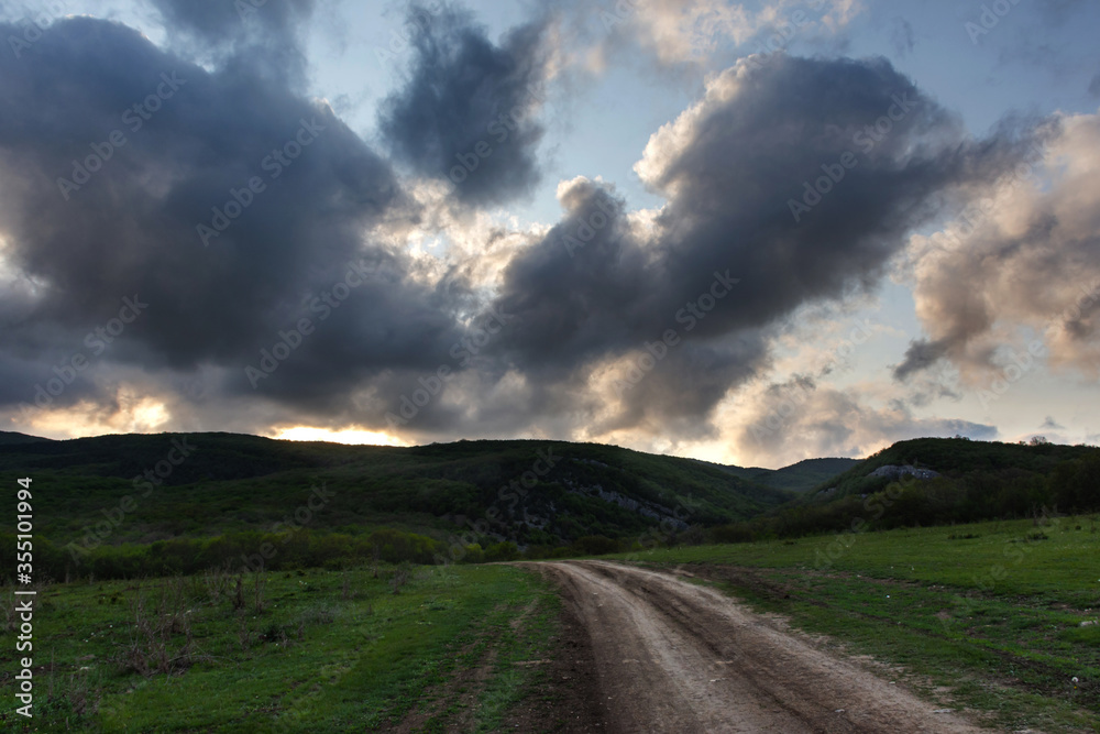 Landscape with a country road and clouds. Threatening rain clouds over the road. The sun breaks through the clouds. Rural spring landscape. A winding path leading into the forest.