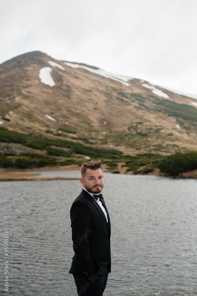 Wedding concept photo shoot. Beautiful young groom standing on a big stone in a lake and mountains with snow Wedding concept photo shoot. Beautiful young groom standing on a big stone in a lake and mountains with snow