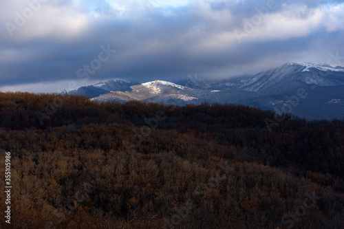 Panorama of snowy mountains on the background of an autumn forest. Snow peaks illuminated by the sun. Storm clouds hang over the mountain. Cold autumn landscape. Crimean mountains in January.