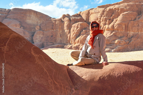 Young woman wearing warm jacket and scarf around head smiling, sitting at naturally formed rock window in wadi rum desert on sunny day