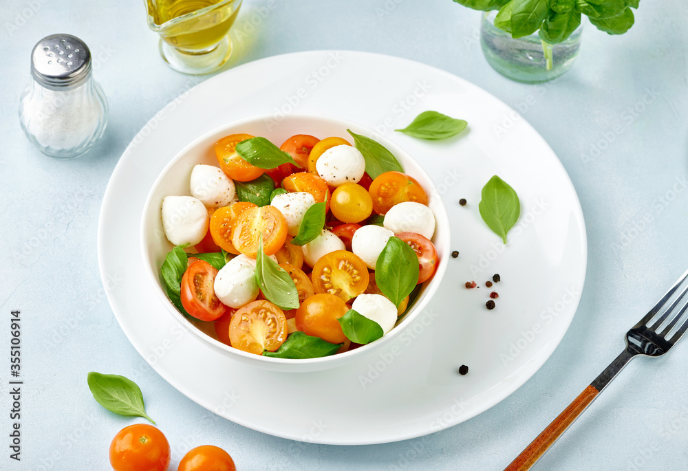 Caprese salad with tomatoes and mini mozzarella cheese in white bowl on white plate. Bocconcini mozzarella cheese with basil leaves. Caprese salad with tomatoes and mini mozzarella cheese in white bowl on white plate. Bocconcini mozzarella cheese with basil leaves.