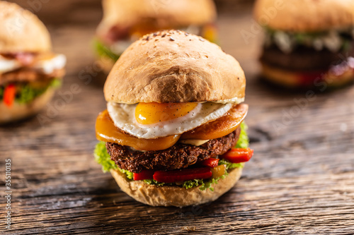 Detail of a hamburger with cheddar, fried egg, red peppers and salad in a crusty bun, with three more burgers in the back