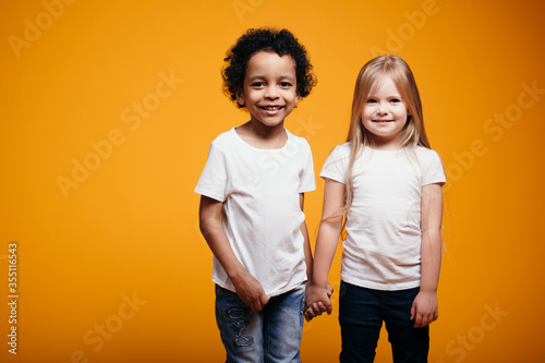 Portrait of two young children in the studio. A dark-skinned boy and a blonde girl are posing for the camera on an orange background in the studio.