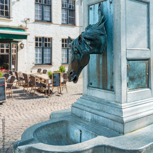 Belgium, Bruges - March 27, 2015 - an antique fountain in the form of a horse's head against the background of a street cafe