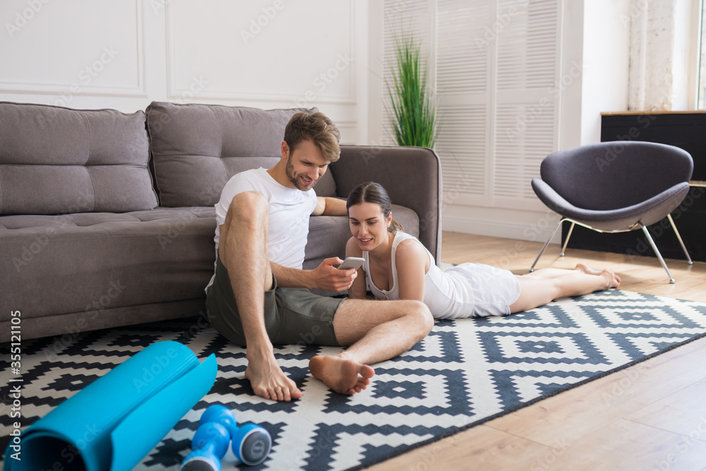 Young couple sitting on the floor after workout and watching something online Young couple sitting on the floor after workout and watching something online